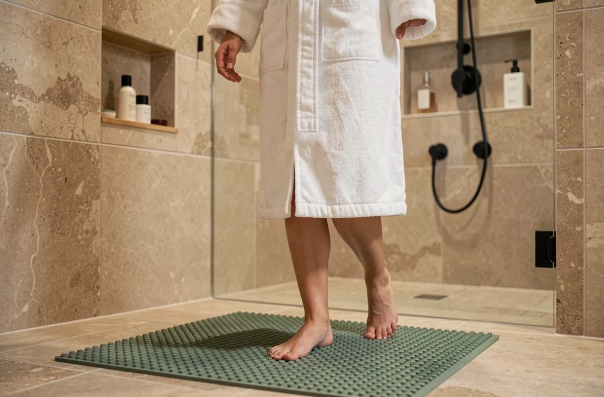 Femme en peignoir blanc posant le pied sur un tapis de bain antidérapant vert sauge devant une douche à l'italienne